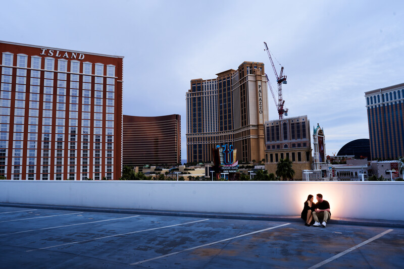 Two silhouetted figures sitting on a blue plaza at dusk with illuminated hotel and casino buildings behind them.
