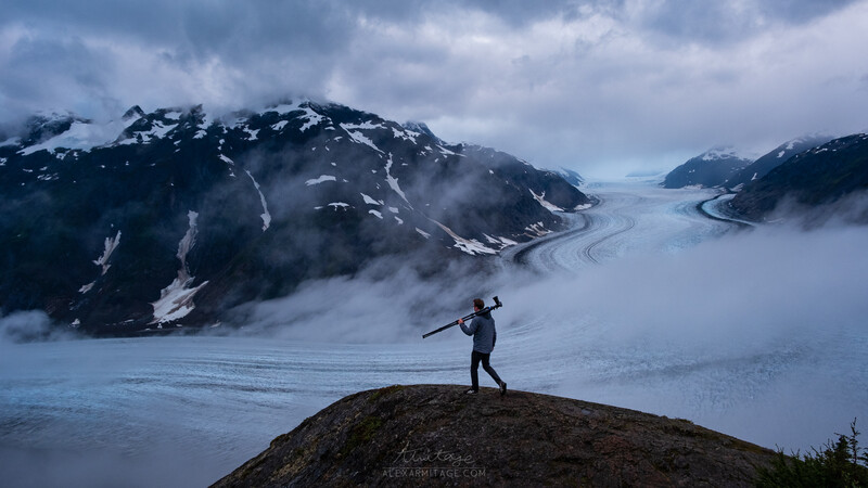 Silhouetted figure standing on a rocky outcrop overlooking a vast glacier valley surrounded by snow-capped mountains.