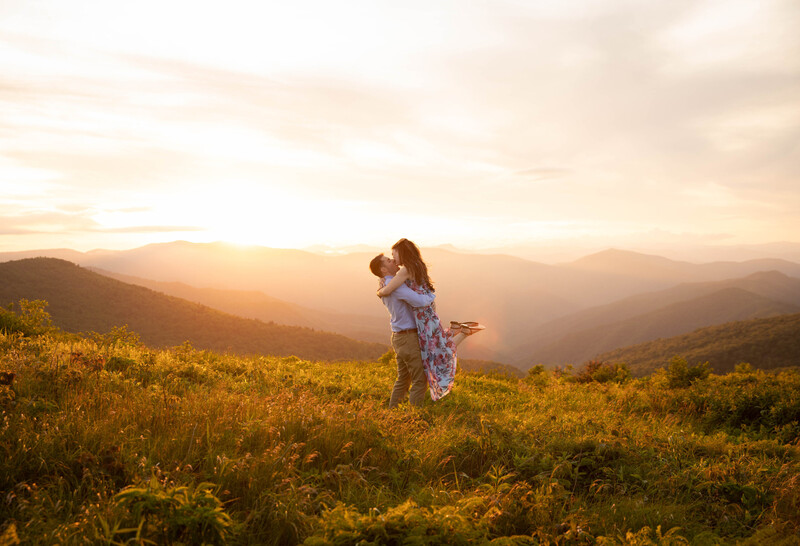 Woman in light clothing standing in a golden wildflower field at sunset, mountains visible in the distance.