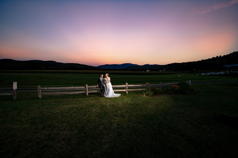 Bride and groom standing together by a wooden fence in a sprawling rural meadow during golden hour.