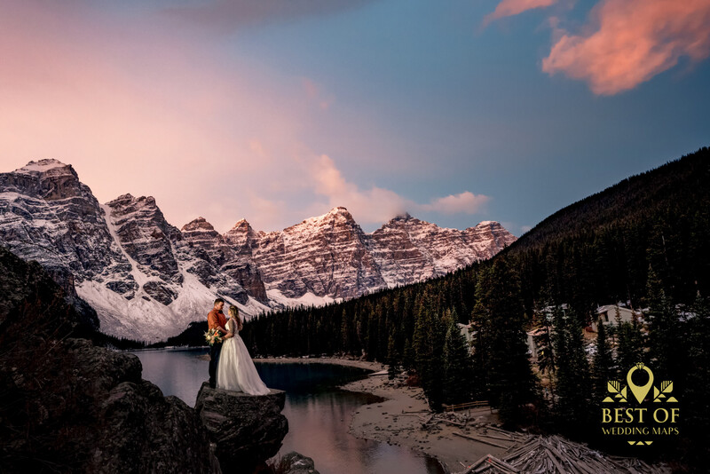Bride in white wedding dress posing on rocky outcrop beside alpine lake with snow-capped mountain peaks and evergreen forest.