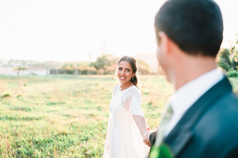 Couple standing in an open field at golden hour, woman in white dress looking back at man in blue suit.