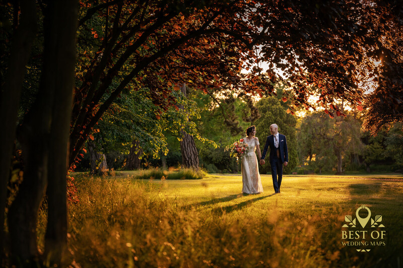 Bride and groom walking together under a canopy of golden-lit trees in a sunlit field.