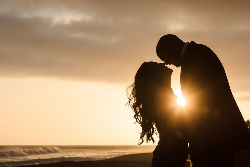Silhouetted couple kissing on a beach at sunset with ocean and dramatic sky in background.