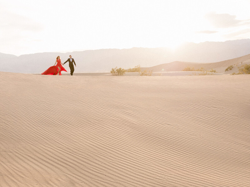 Couple in formal wear walking across desert sand dunes at golden hour with mountains in background.