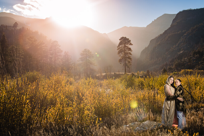 Family of three standing in golden meadow at sunset with forested mountains in background.