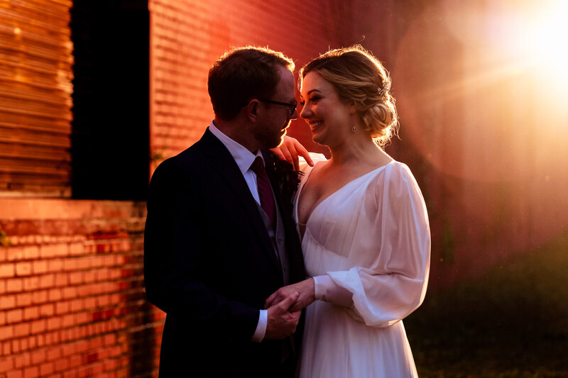 Couple in formal attire facing each other against a brick wall lit with warm golden light.