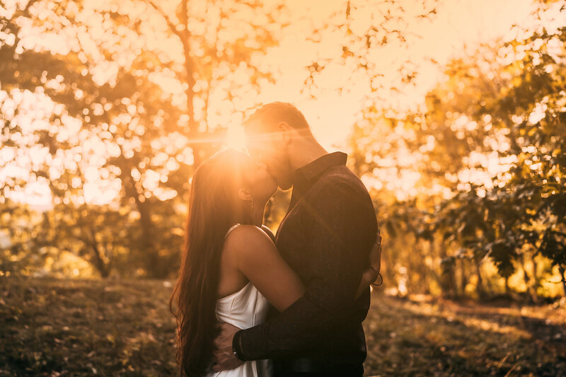 Couple kissing during golden hour with backlit sunlight streaming through trees.