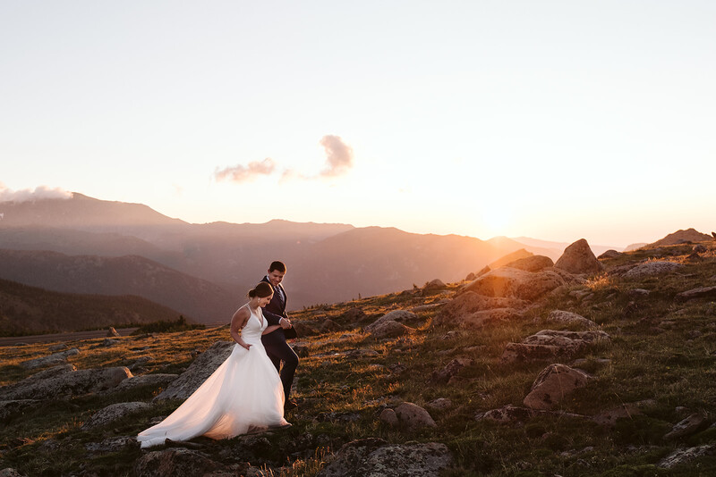 Bride and groom embracing on a rocky hillside at golden hour with mountains in the background.