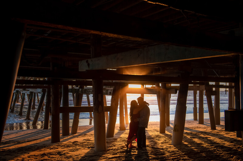 Silhouetted figure standing on sandy beach under wooden pier with golden-hour sunlight streaming through structural supports.
