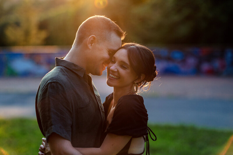 Couple embracing and smiling outdoors during golden hour with stadium seating visible behind them.