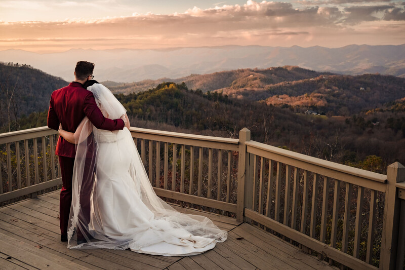 Bride and groom embracing on a wooden deck railing, overlooking a mountainous valley at sunset.