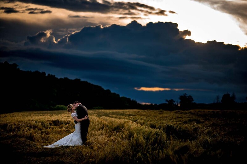 Bride in white dress standing alone in a grassy field beneath a dramatic cloudy sky at dusk.