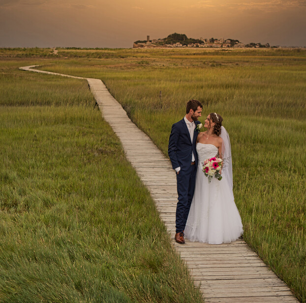 Bride and groom standing together on a wooden boardwalk through a vast marsh landscape at golden hour.