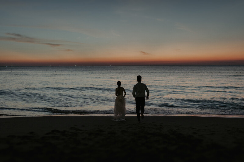 Two figures in formal attire standing at the water's edge at dusk, silhouetted against an orange and purple sunset sky.