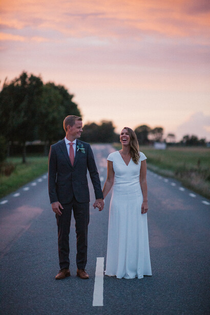 Bride and groom holding hands standing on a rural road at sunset.