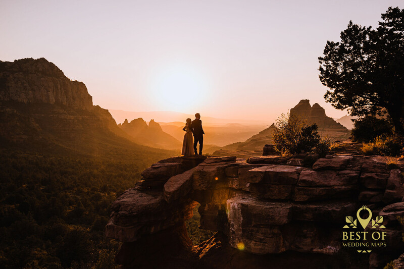 Two silhouetted figures standing on a rocky outcrop at sunset, surrounded by dramatic mountain peaks bathed in golden light.
