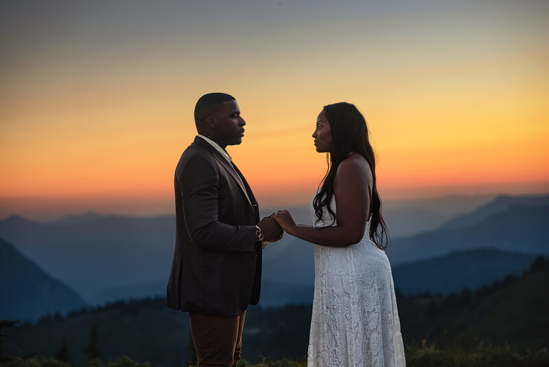 Couple in formal attire holding hands during golden hour with layered mountains in silhouette behind them.