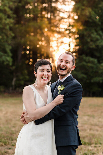Bride and groom laughing together outdoors during golden hour with trees in background.