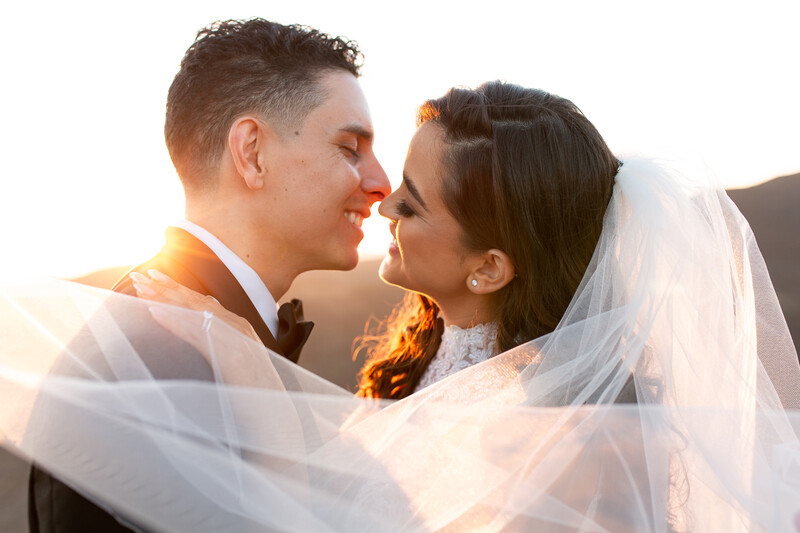 Bride and groom embracing nose-to-nose with backlit golden hour lighting and flowing white veil.