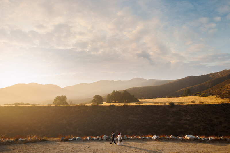 Couple standing in a vast plowed field at golden hour with mountains and valley in the background.
