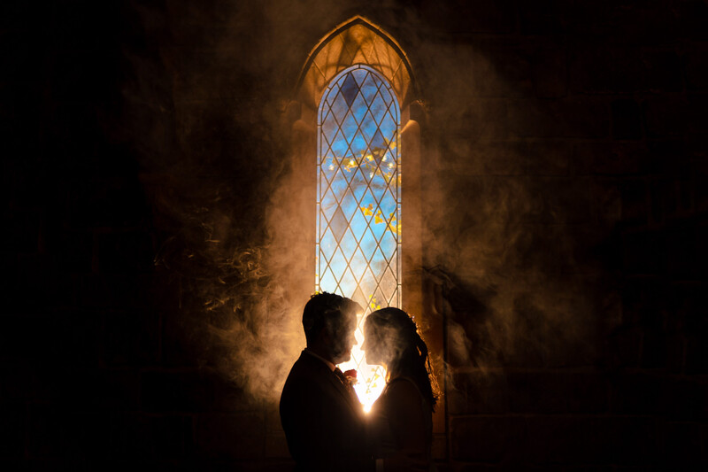 Two silhouetted figures facing each other in front of a tall arched stained glass window backlit with warm golden light.
