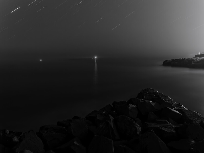 Long exposure photograph of a rocky shoreline at night with calm water and distant lights reflecting on the surface.
