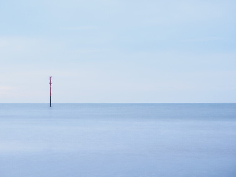 Solitary navigation marker standing in calm, misty water under overcast sky.
