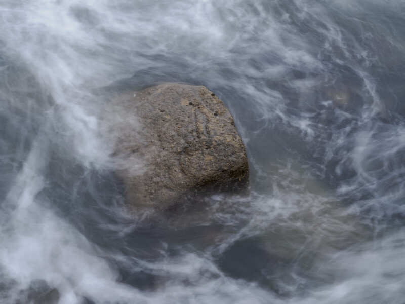 Large boulder surrounded by swirling white foam and turbulent seawater.