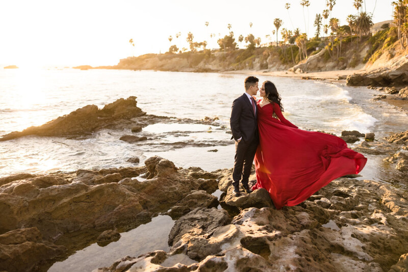 Couple in formal attire kissing on rocky coastline at sunset, woman wearing flowing red dress.