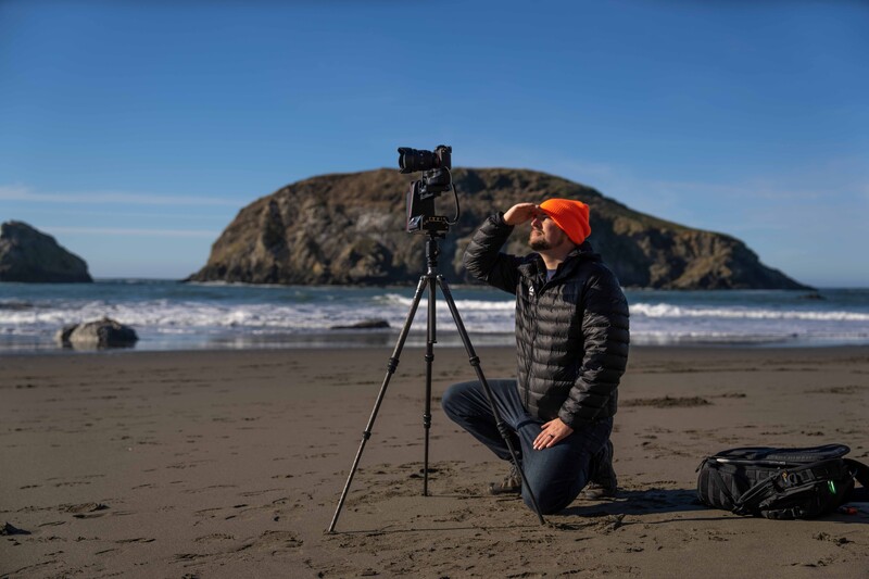 Photographer in orange cap and black jacket crouching beside camera on tripod on a sandy beach, with a large rocky island and waves in the background.