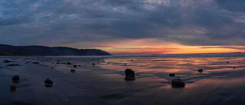 Rocky beach at sunset with dark boulders scattered across wet sand and calm water reflecting golden and blue twilight sky.