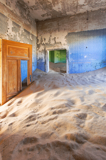 Abandoned building interior filled with sand, featuring blue painted walls and an orange wooden door.