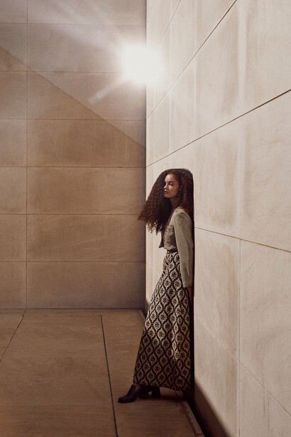Woman with long dark hair leaning against beige tile wall in narrow corridor with bright overhead lighting.