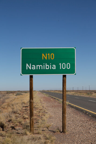 Green highway sign reading N10 and Namibia 100 mounted on wooden posts beside a desert road.