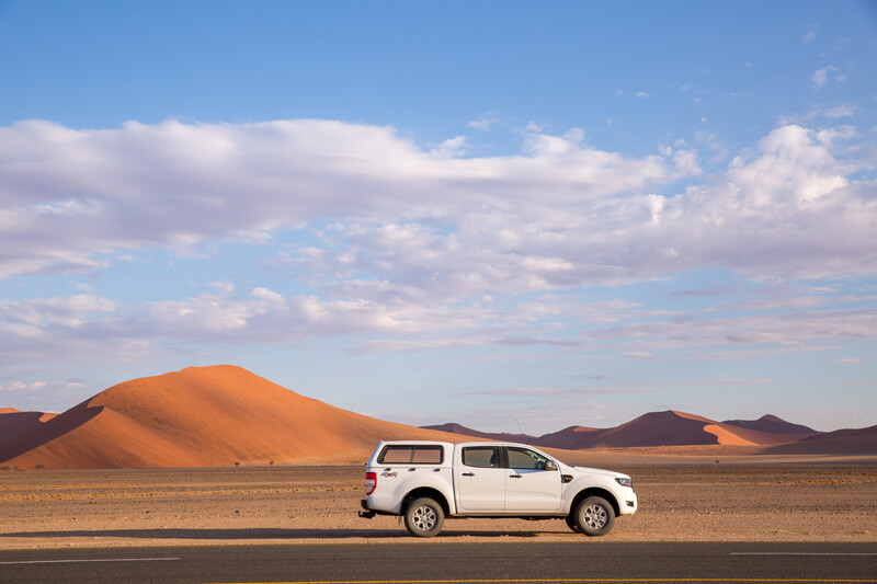 White pickup truck parked on a desert highway with large sand dunes in the background under a partly cloudy sky.