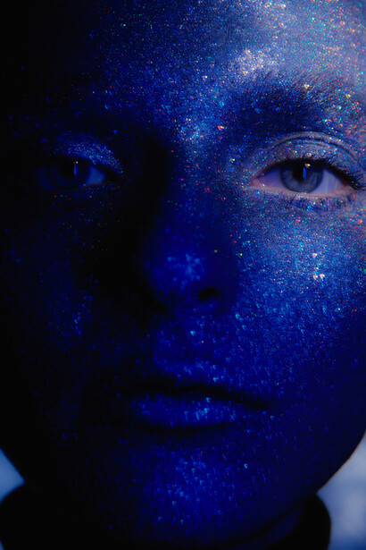 Person's face covered in blue glitter and sparkles against a dark background, with one eye visible and direct gaze.