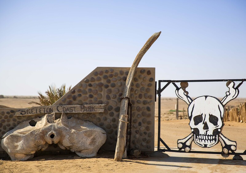 Desert roadside attraction with hippo sculpture, wooden post, and skull-and-crossbones metal sign.