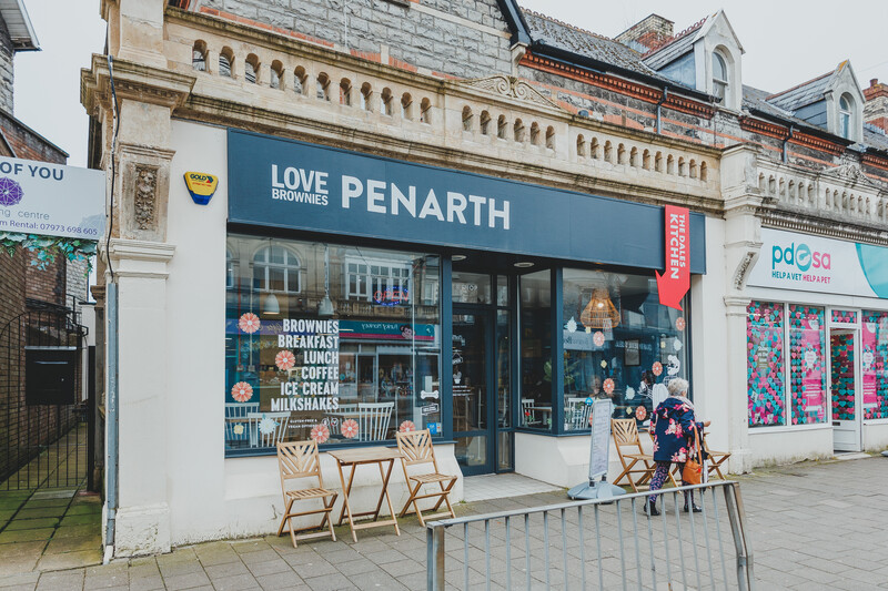 Storefront of Love Brownies Penarth with blue signage, display windows, and wooden deck chairs on the pavement.