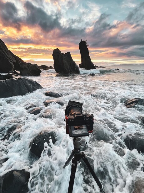 Camera on tripod positioned in crashing waves with sea stacks silhouetted against dramatic sunset sky.