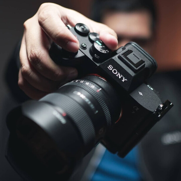 Photographer holding Sony mirrorless camera with attached lens, hands gripping body and grip area.