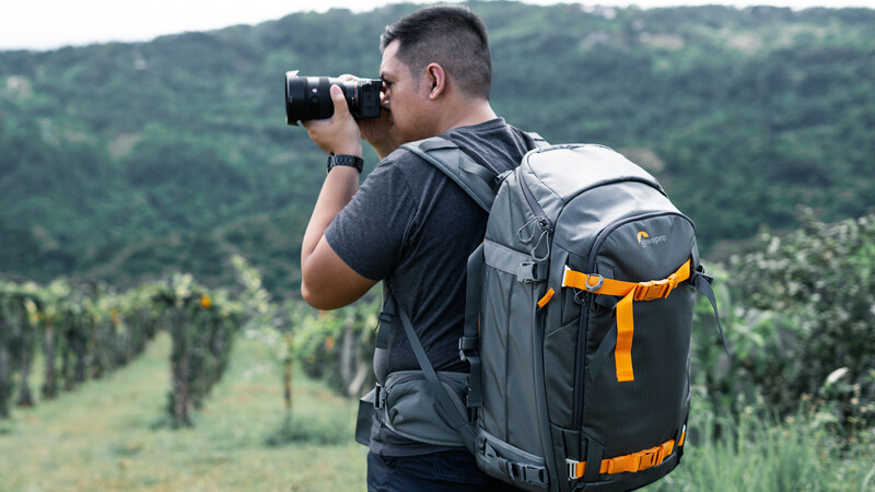 Photographer with backpack holding camera to face while photographing terraced vineyard landscape.