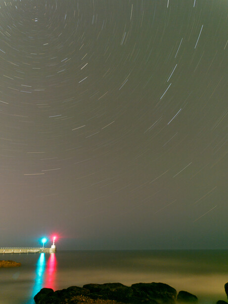 Long-exposure photograph of star trails above a dark coastline, illuminated by red and blue lighthouse beams.