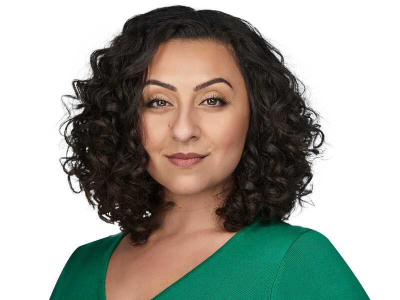 Woman with shoulder-length curly dark hair wearing a green v-neck top against a white background.