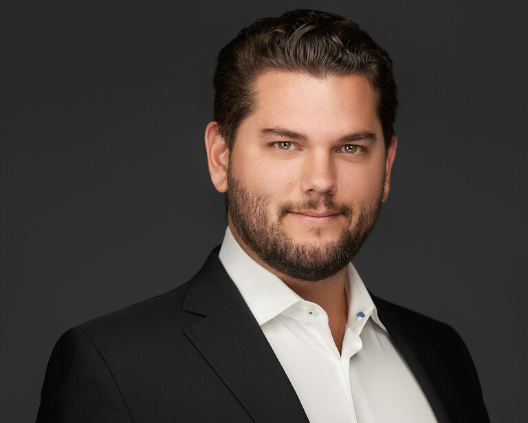 Professional headshot of a man in a black blazer and white shirt against a neutral gray background.