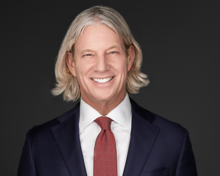 Professional headshot of a man with shoulder-length blonde hair wearing a dark navy suit and red tie.