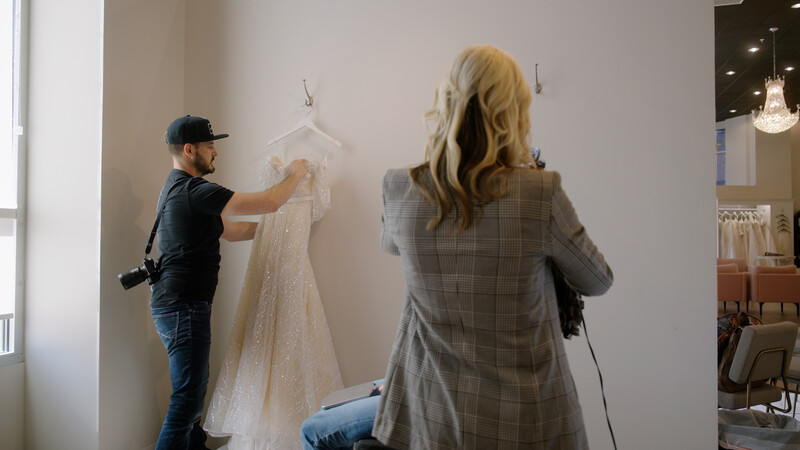Photographer and assistant reviewing a delicate lace wedding dress displayed on a hanger against a studio wall.