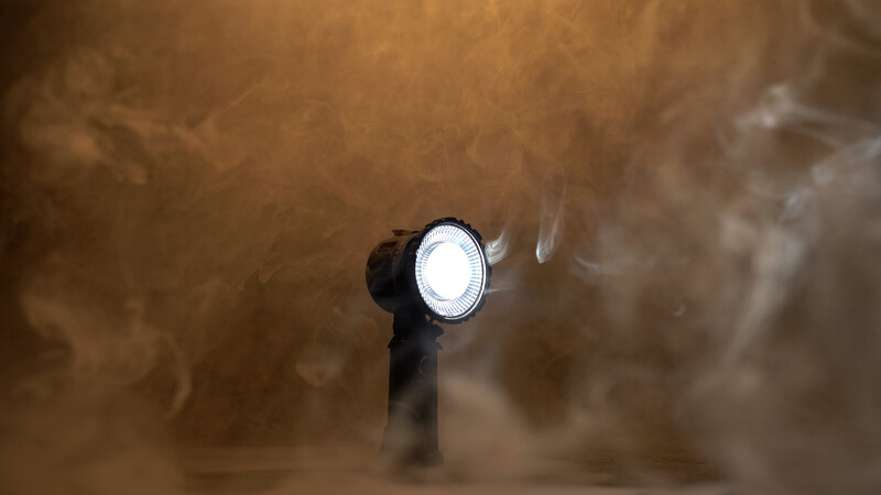 Handheld spotlight with bright white LED beam cutting through thick brown dust or smoke cloud.