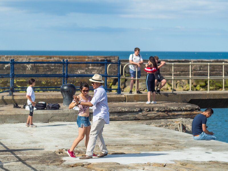 An adult in white clothing and hat dances with a child on a waterfront promenade while others watch nearby.