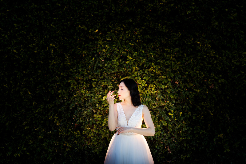 Woman in white dress eating from a plate against a dark green foliage backdrop.